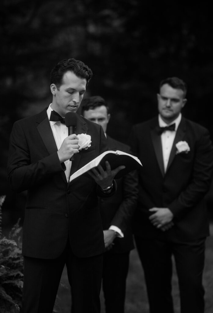 Groomsman reading from a Bible during an outdoor wedding ceremony, wearing a black tuxedo and bow tie, captured in black and white for a timeless, emotional moment.