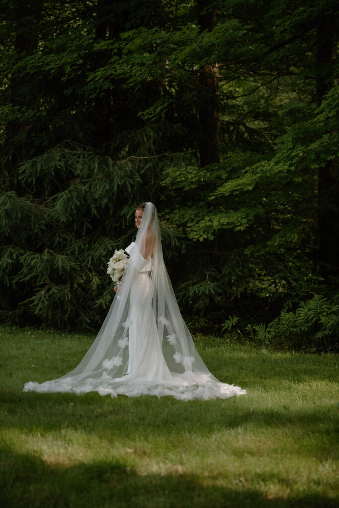 Bride standing in a forest clearing wearing a fitted wedding gown and cathedral veil, holding a bouquet of white flowers, captured in soft, natural light.