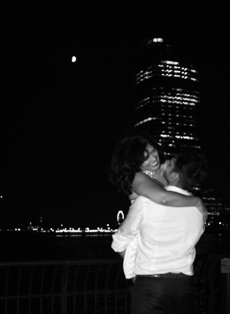 Man lifts his fiancée under the moonlight with the city skyline glowing in the background, captured with dramatic flash.