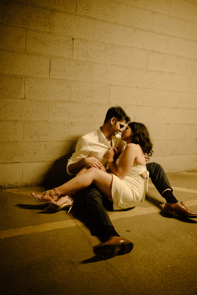 Couple seated on the ground in a passionate embrace, lit by flash against a concrete wall during a nighttime session.