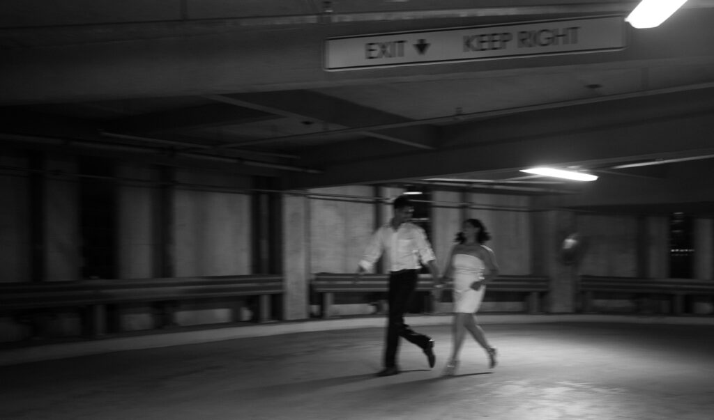 Blurry motion flash shot of couple holding hands and running through a dimly lit parking garage, capturing the thrill of the night.