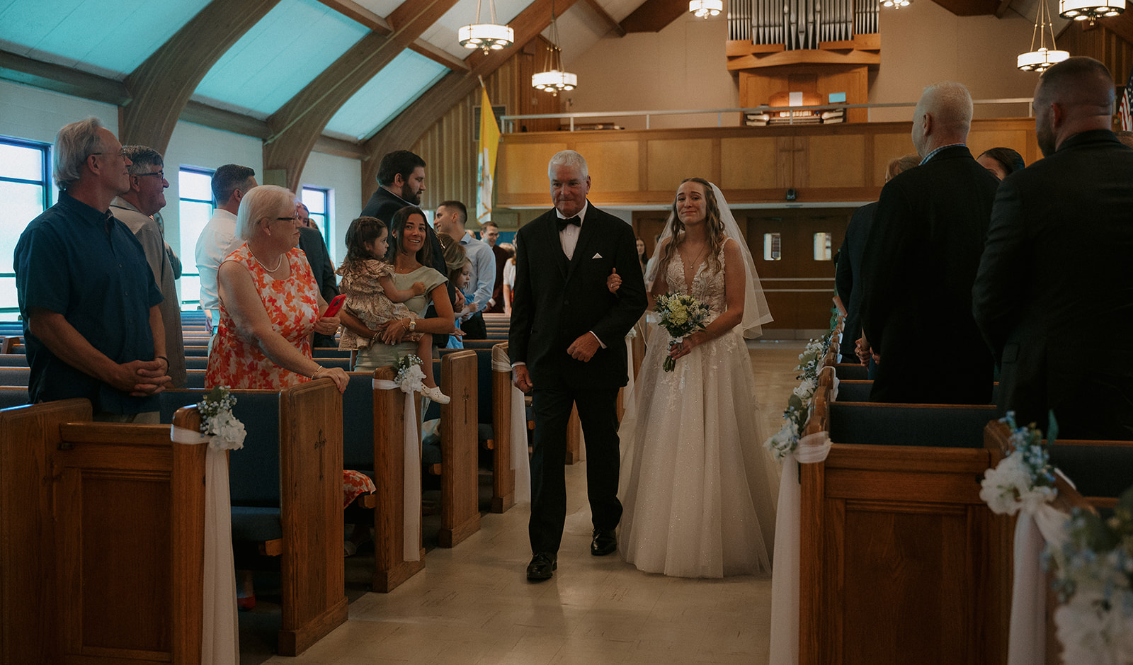 Bride walking down aisle with her father during Catholic wedding ceremony, guests smiling — storytelling wedding photography moment.