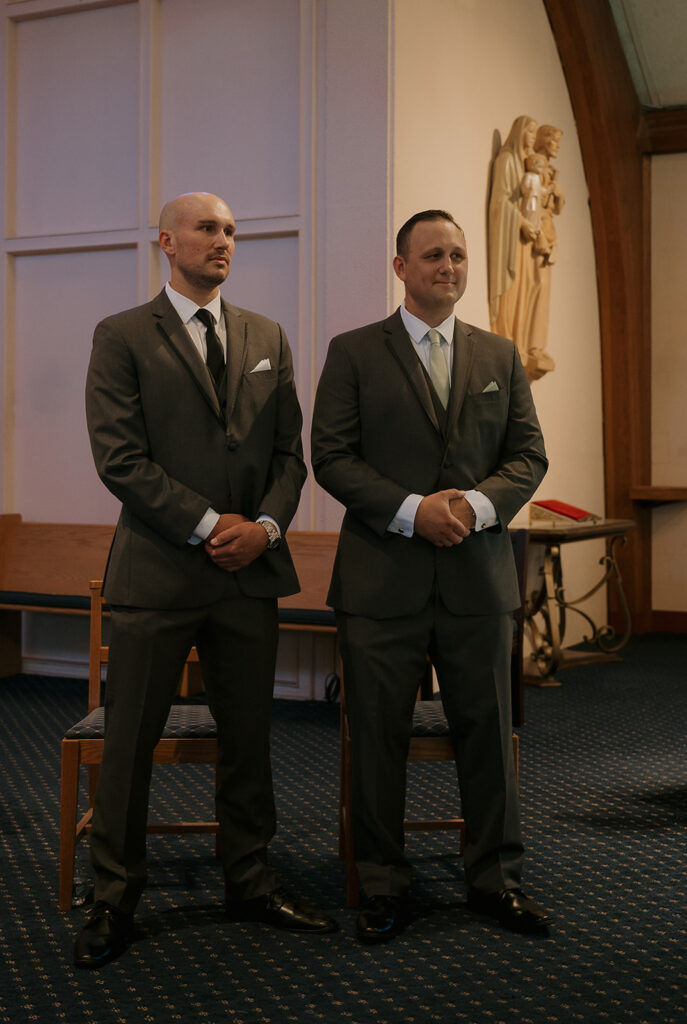 Groom standing at altar with best man, watching bride enter the church — storytelling wedding photography candid.