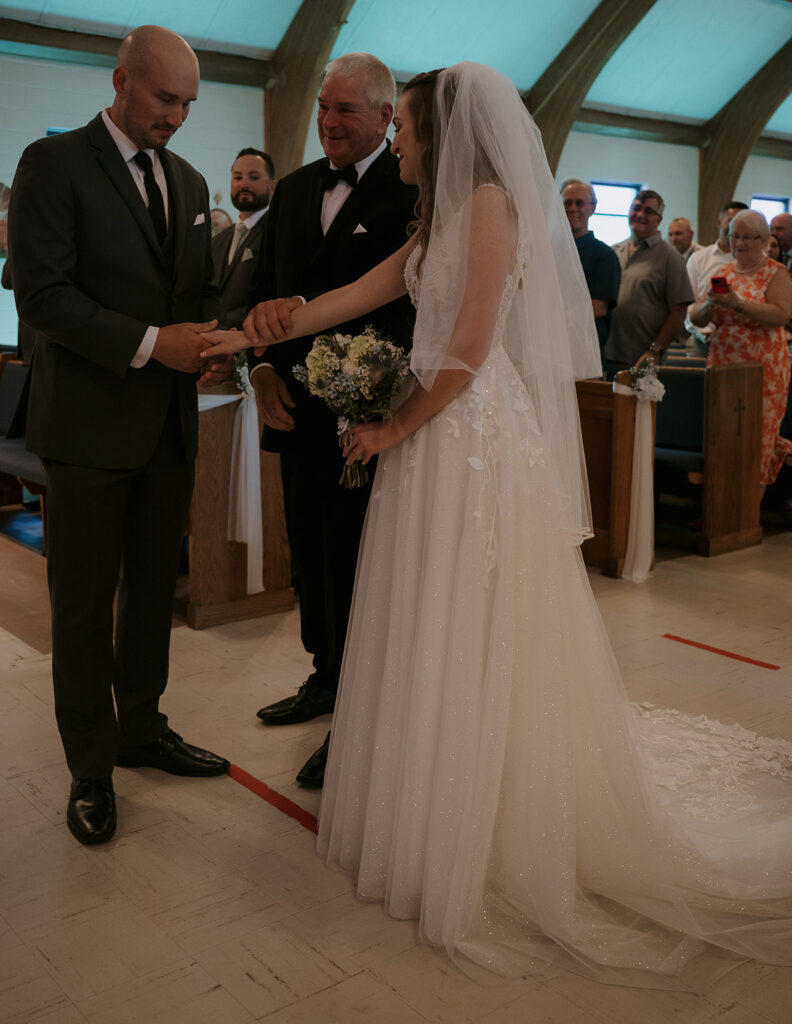 Bride’s father placing her hand into groom’s during Catholic ceremony — storytelling wedding photography of giving away.