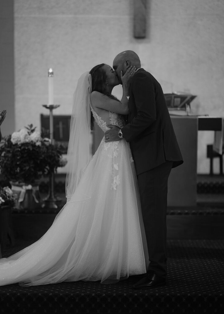 Bride and groom sharing first kiss at Catholic altar — storytelling wedding photography highlight moment.