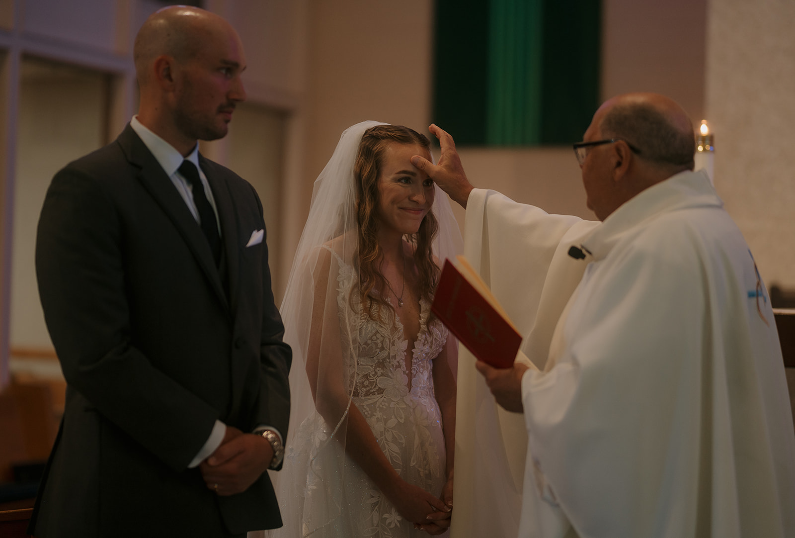 Priest blessing bride during Catholic ceremony, groom standing beside — storytelling wedding photography faith-centered detail.