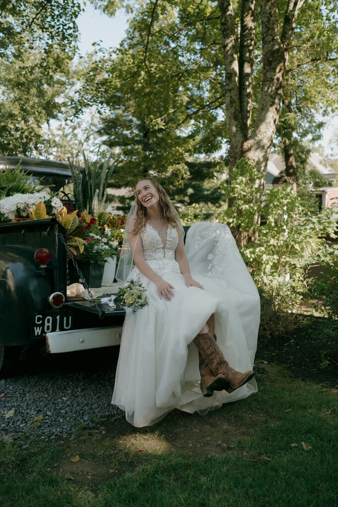 Bride sitting on vintage truck bed filled with flowers, wearing cowboy boots — storytelling wedding photography at The Inn at Millrace Pond.