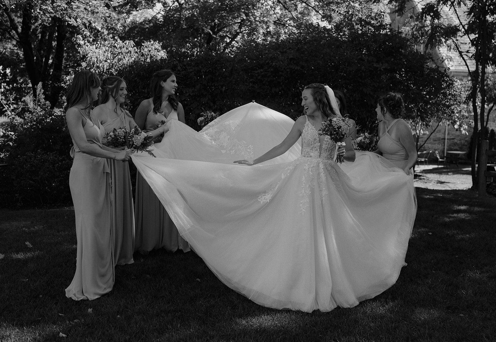 Bride surrounded by bridesmaids holding out her flowing gown, joyful portraits at The Inn at Millrace Pond.