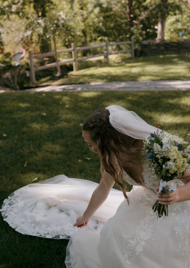Bride adjusting her dress on the grass, bouquet in hand — candid storytelling wedding photography at The Inn at Millrace Pond.