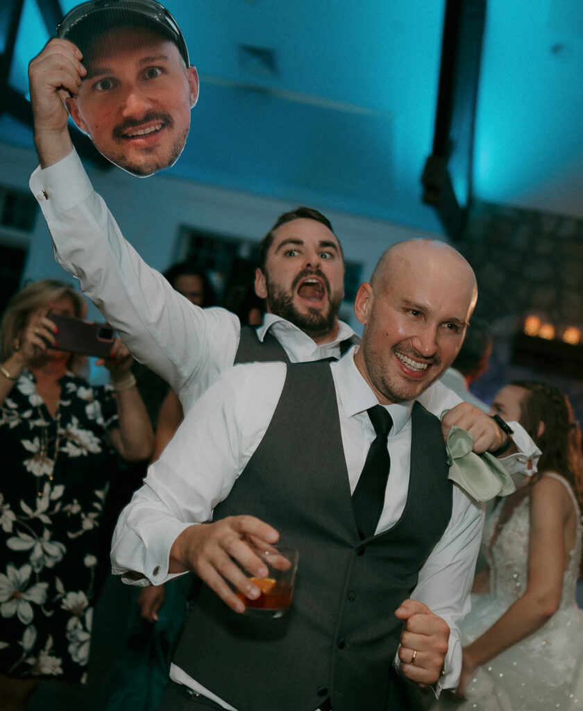 Groom smiling with friends as guest waves oversized cutout head on dance floor at The Inn at Millrace Pond wedding reception.
