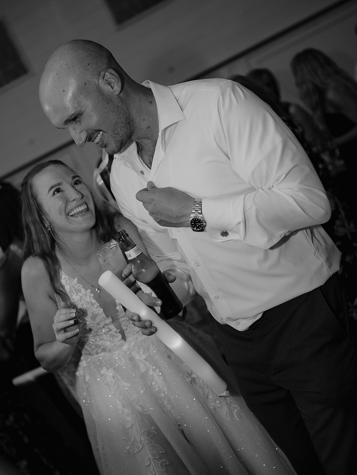 Bride and groom laughing together while holding drinks and glow sticks during barn wedding reception at The Inn at Millrace Pond.