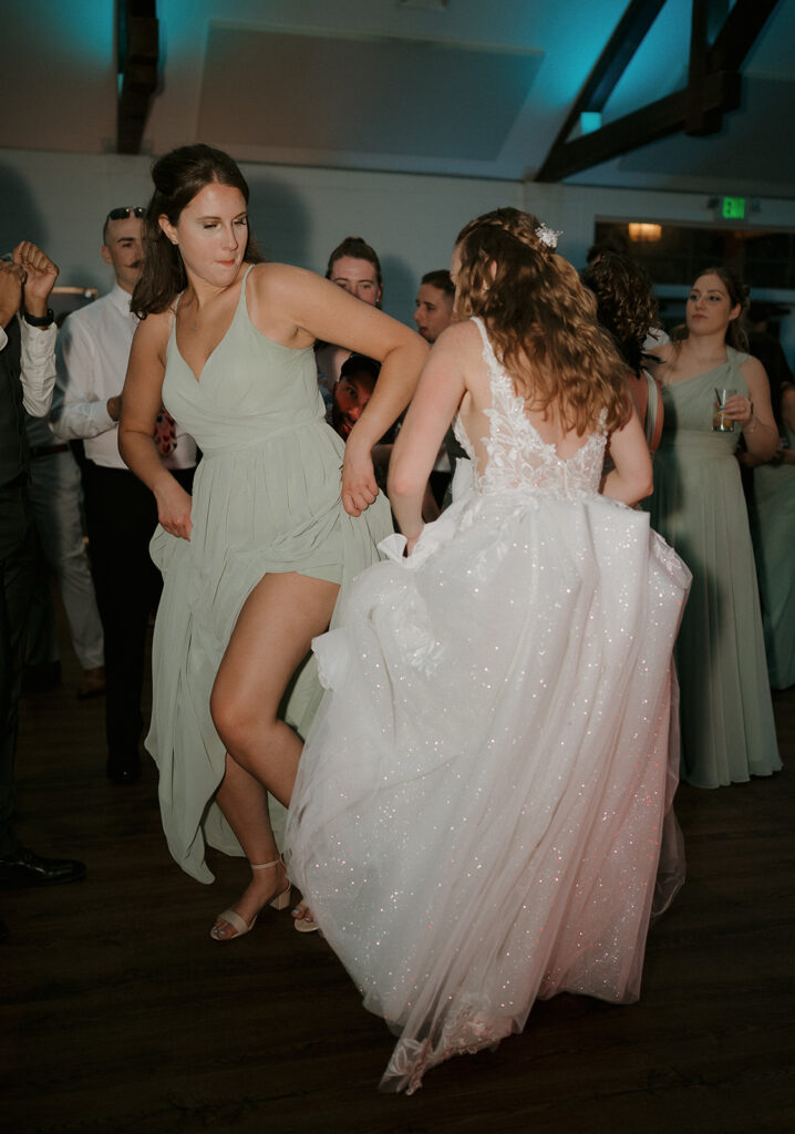 Bride dancing with bridesmaid in sage green dress, candid moment at The Inn at Millrace Pond barn reception.