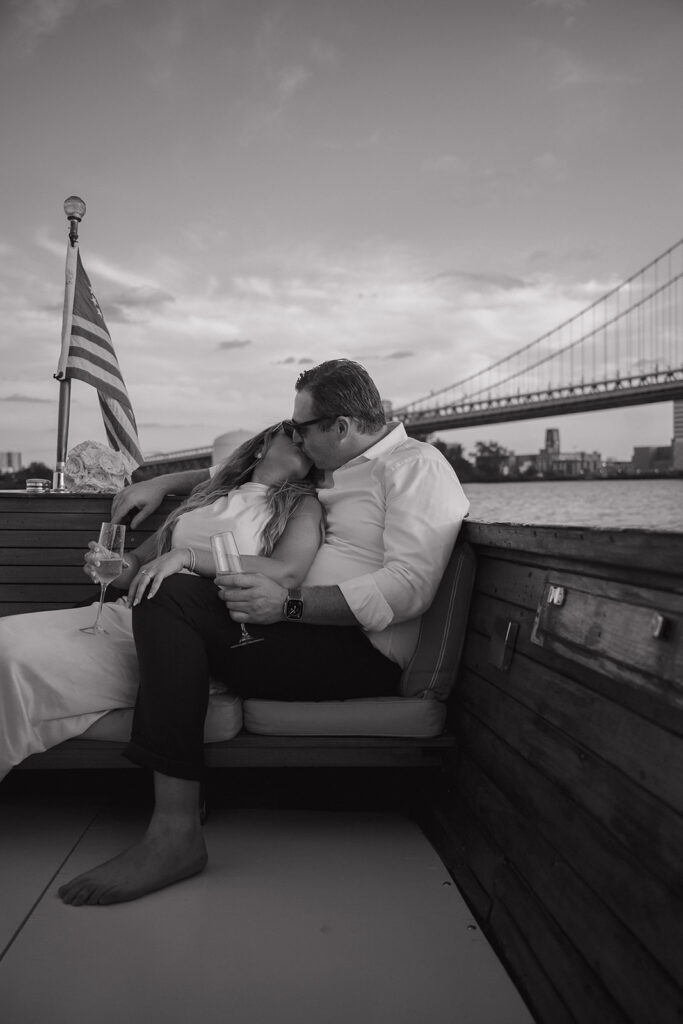 Black and white photo of a couple sharing a kiss with champagne glasses on The Argonaut during their dreamy engagement photos in Philadelphia.