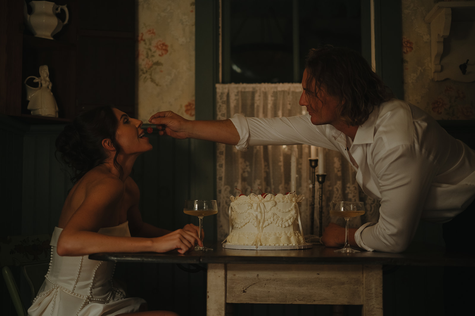 Groom feeding bride cherries over a vintage cake during their playful, late-night intimate wedding session photographed in Frenchtown, NJ.