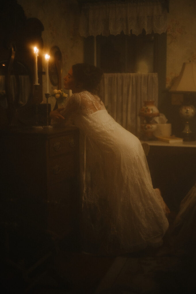 Bride in a lace robe leaning toward her vanity in soft candlelight, creating romantic and nostalgic imagery typical of Ell Peters’ intimate wedding photos.
