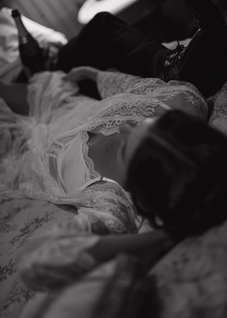 A dreamy black-and-white shot of the bride resting on the bed in lace, champagne nearby, creating a moody and cinematic portrait by Ell Peters.