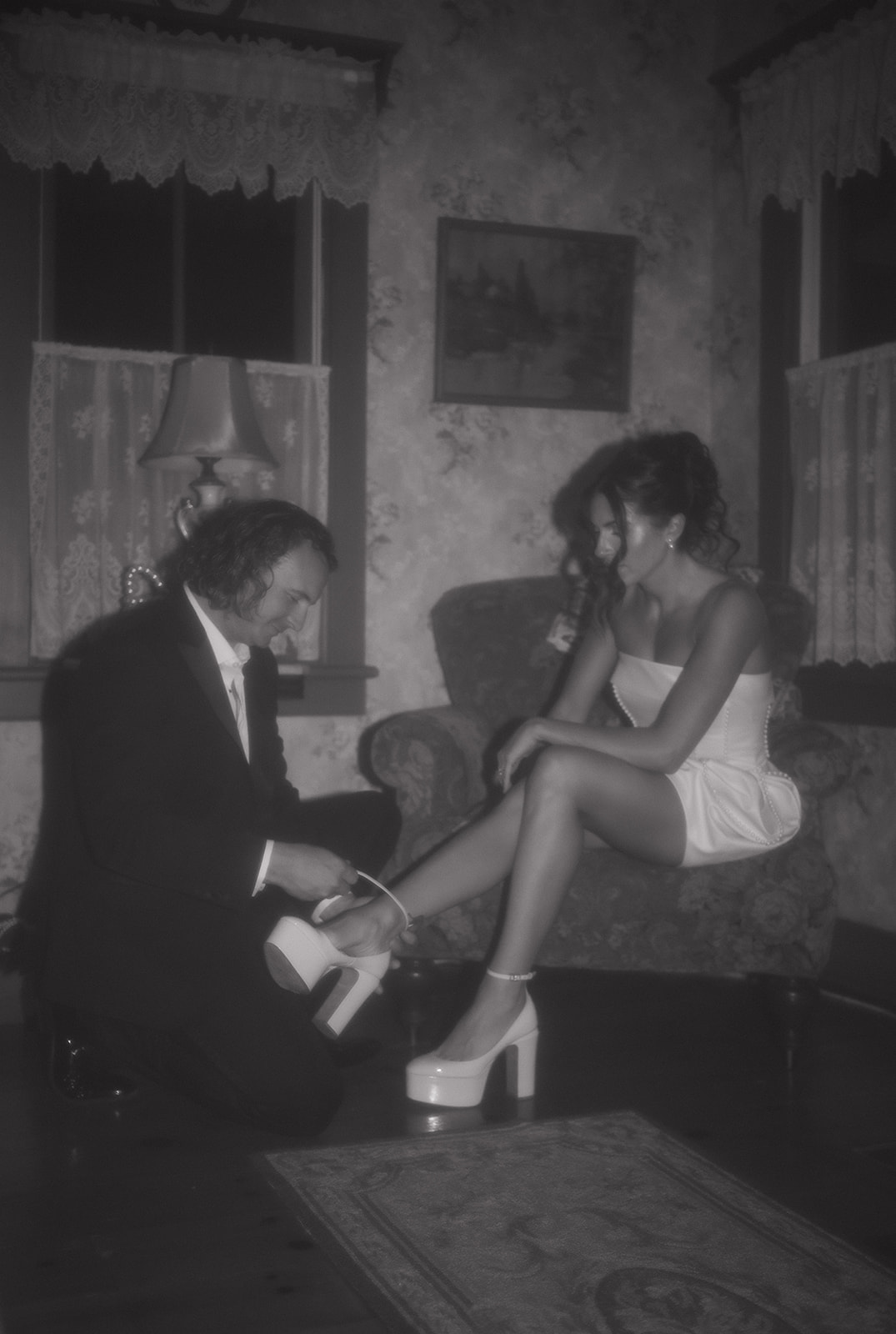 A cinematic black-and-white moment of the groom kneeling to fasten the bride’s heel during their late-night intimate wedding photos at River Witch Cottage in New Jersey.