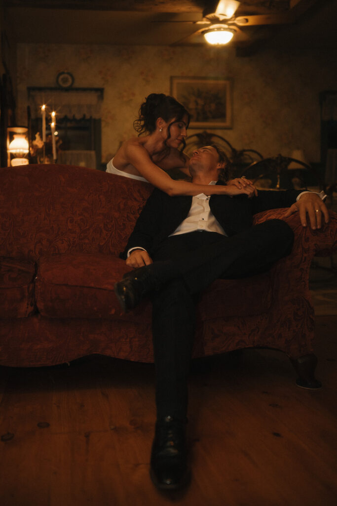 The bride leans over the couch to cradle the groom’s face in soft candlelight, captured in a warm, moody style by New Jersey wedding photographer Ell Peters.