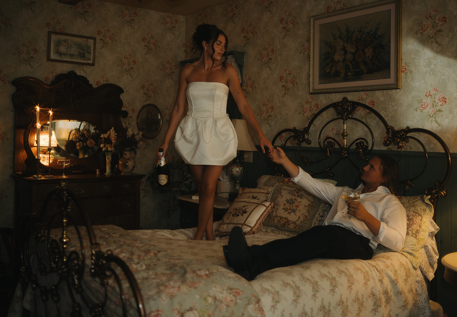 The bride stands barefoot on the bed holding champagne, reaching for her groom in a playful, romantic moment during their intimate New Jersey wedding photo session.