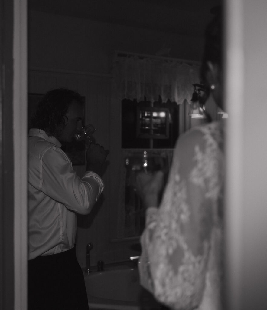 A black-and-white candid of the groom sipping wine in the vintage bathroom while the bride stands in soft focus, adding a documentary feel to their intimate wedding photos.