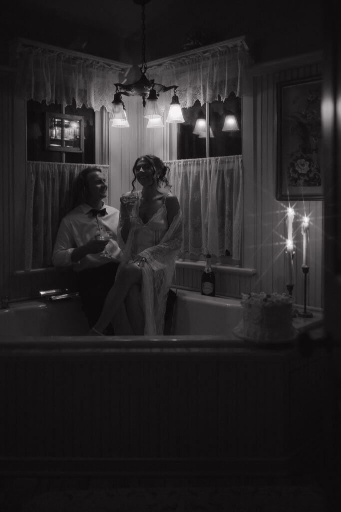 The couple sits together in the corner bathtub, laughing and holding drinks under soft bathroom lights, creating a cozy late-night moment in their New Jersey wedding portraits.
