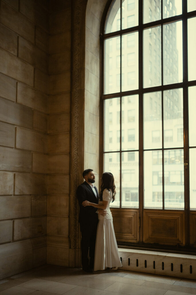Couple holding each other by a large window for their New York Public Library engagement photos.
