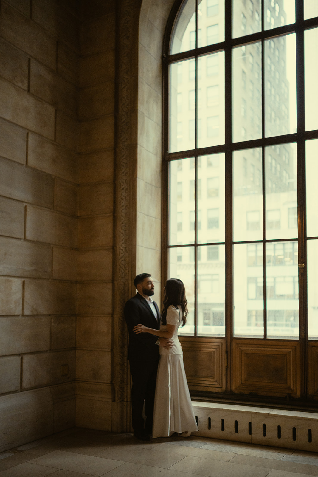 Couple holding each other by a large window for their New York Public Library engagement photos.