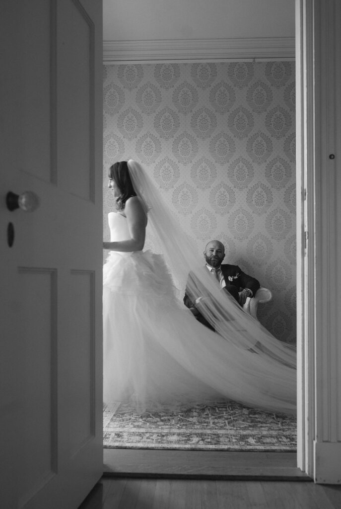 Black and white portrait of bride walking past doorway with veil trailing inside Stone Acres Farm manor house.