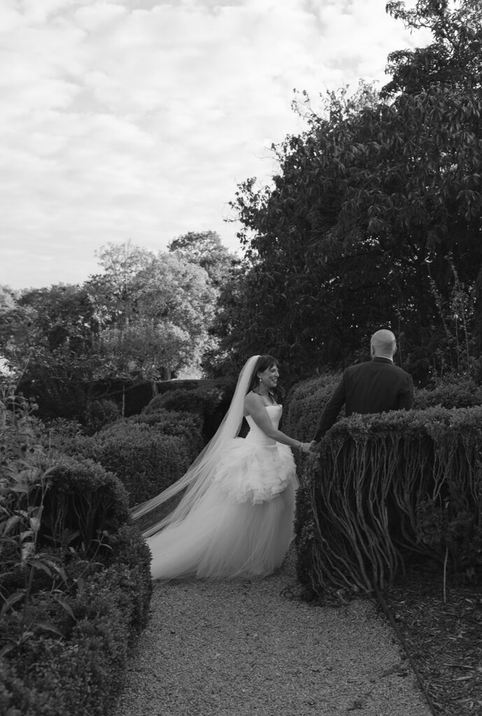 Black and white photo of a bride and groom walking through the Boxwood Garden at Stone Acres Farm.