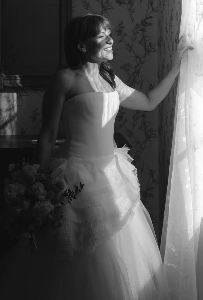 Black and white photo of a bride in Vera Wang gown holding bouquet by window in soft morning light during Stone Acres Farm wedding getting ready moments.