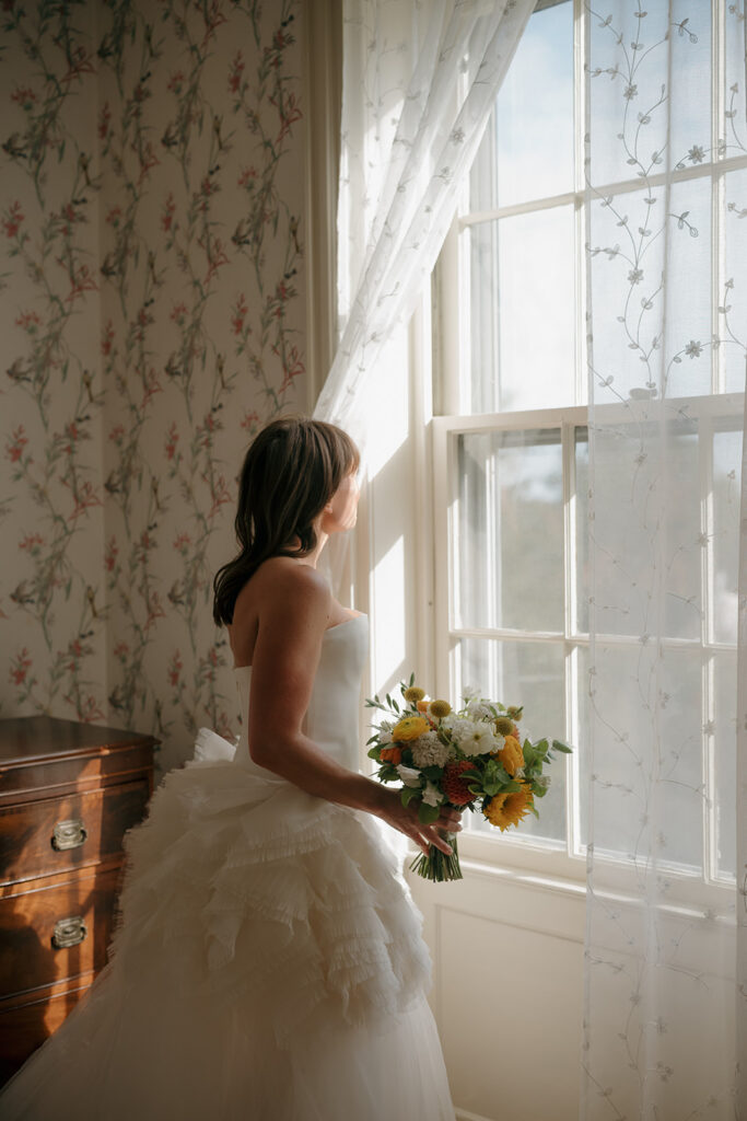 Bride in Vera Wang gown holding bouquet by window in soft morning light during Stone Acres Farm wedding getting ready moments.