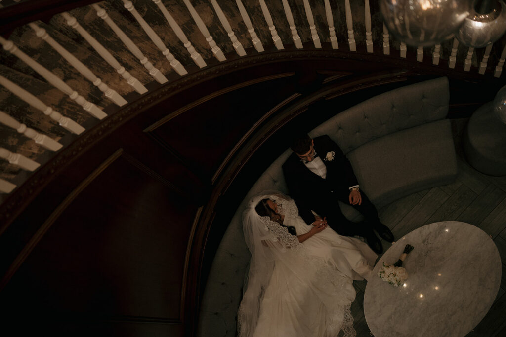 Cinematic wedding photography of bride and groom sitting together on a curved staircase couch in a moody interior.