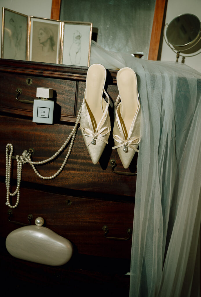 Flash shot of a brides wedding details styled on a dresser with heels, pearls, perfume, and veil. 