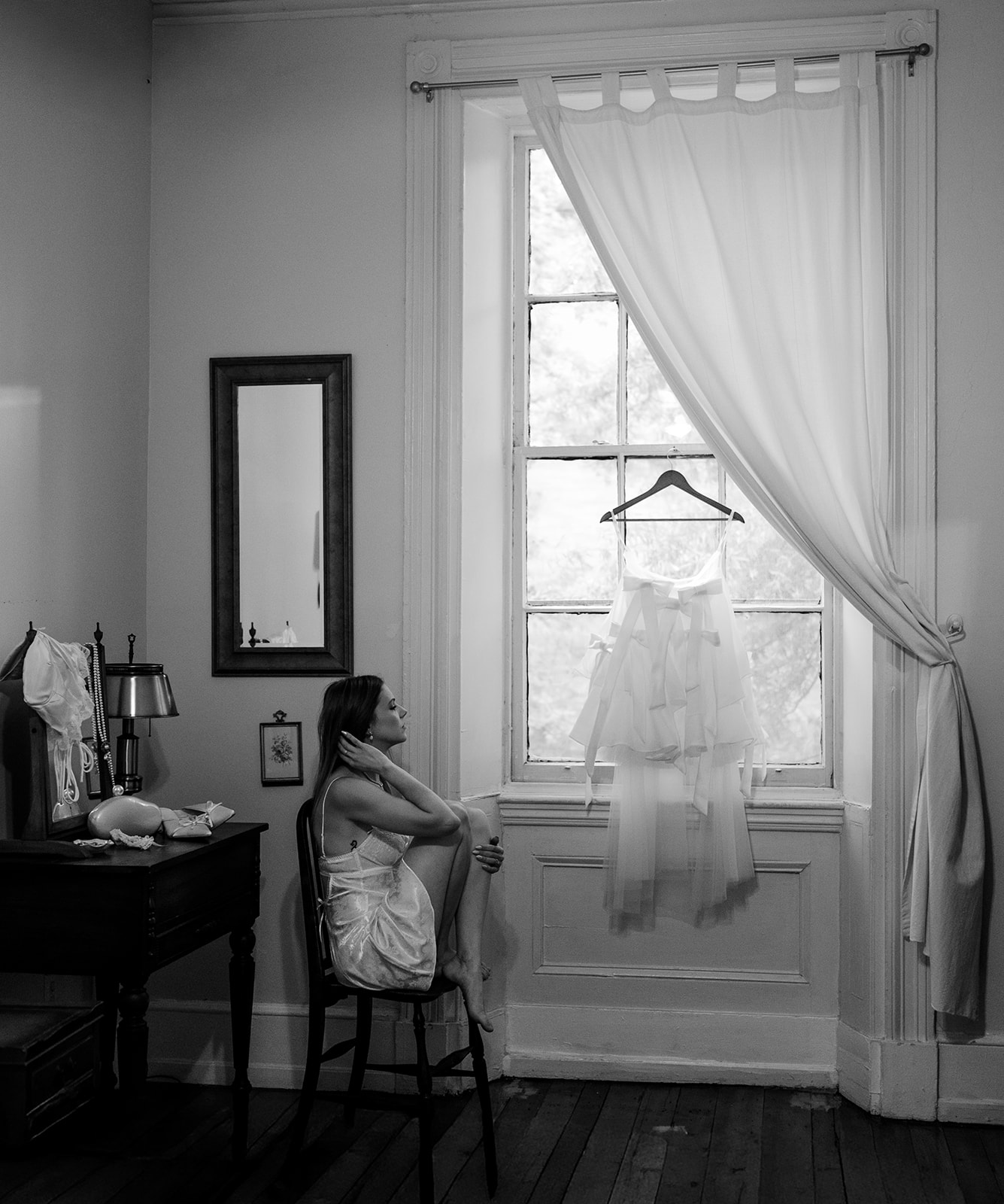 Black and white photo of a bride posing by a window during her getting ready wedding photos with her dress hanging on the window at Vaux Studio in Philadelphia.