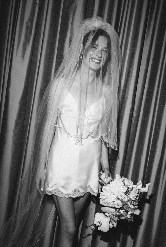 Black and white flash photo of a bride wearing a robe and her veil while holding her bouquet. 