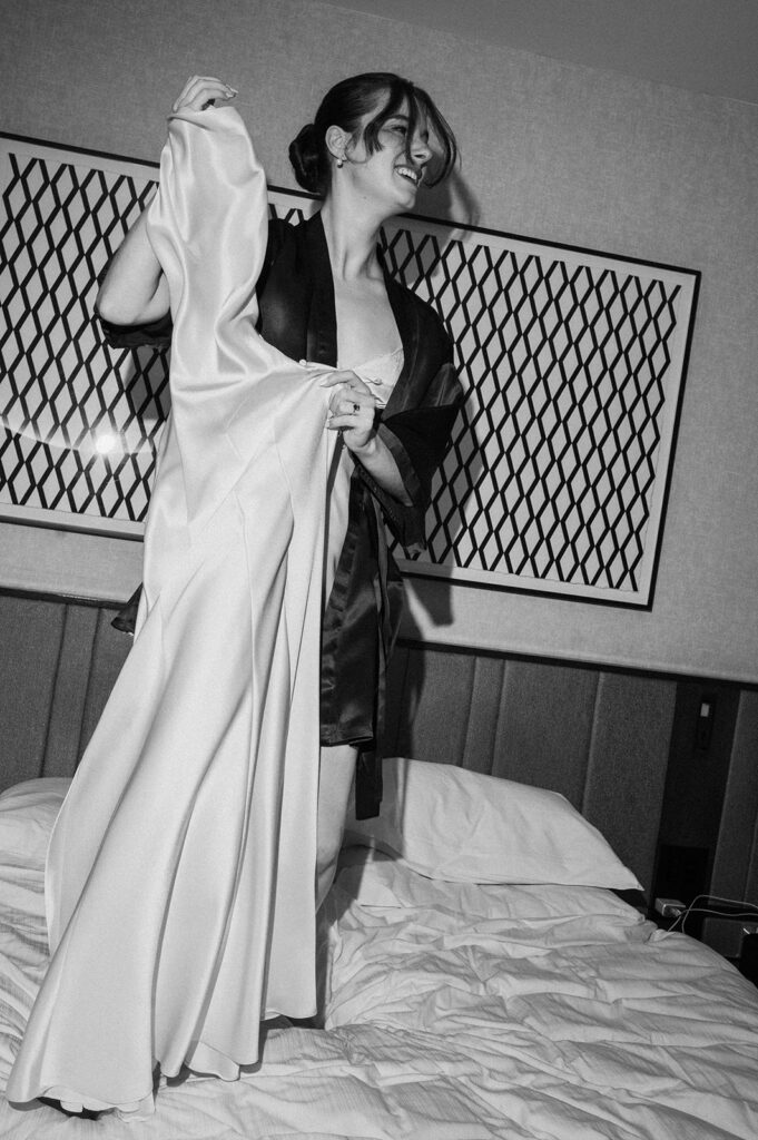 Black and white flash photo of a bride standing om her hotel bed with her dress in hand. 
