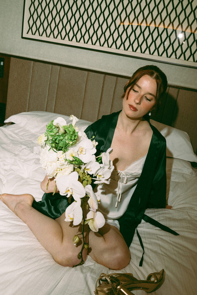Bride sitting on her hotel bed in her rove with her bouquet and heels. 