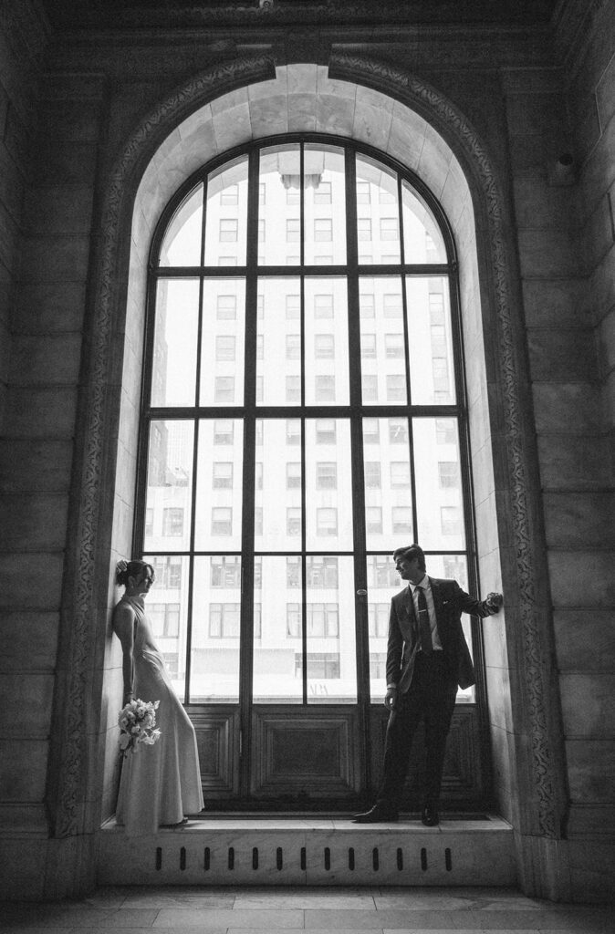 Black and white photo of a bride and groom posing in a window at The New York Public Library. 