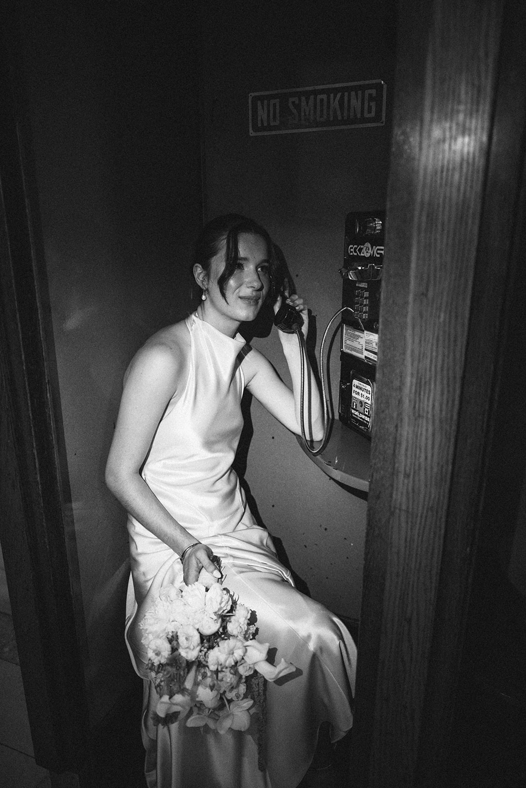 Black and white photo of a bride sitting at a telephone booth. 